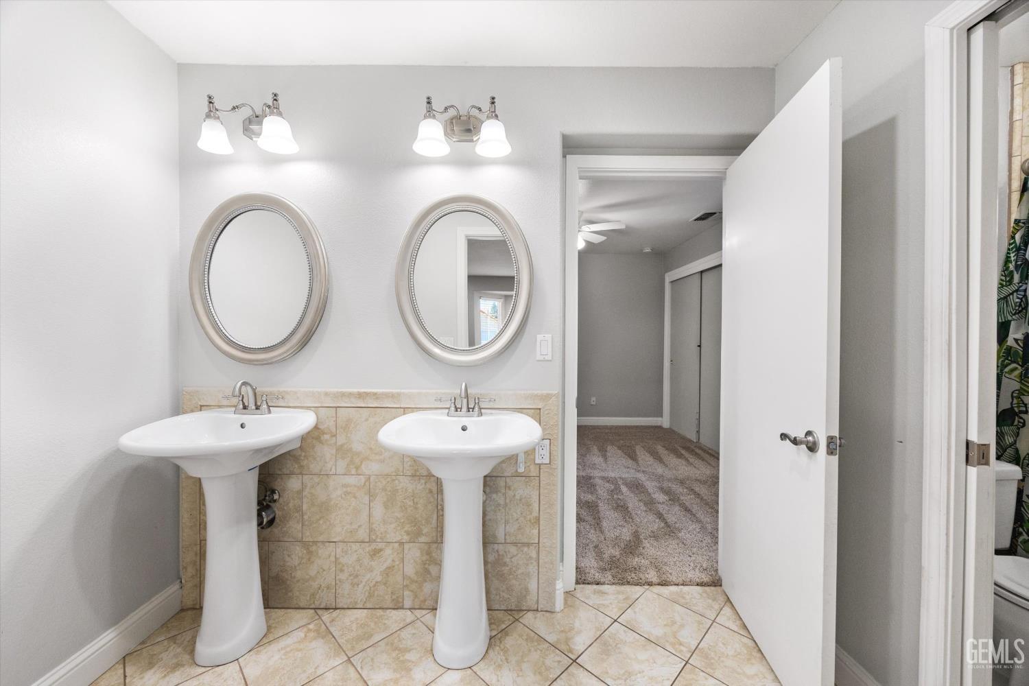 Bathroom with two pedestal sinks and oval mirrors beside an open door to a carpeted room.