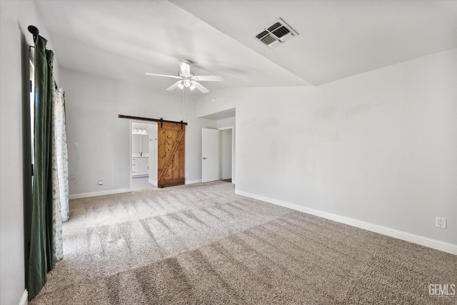 Empty carpeted bedroom with a vaulted ceiling, ceiling fan, and wooden sliding barn door.