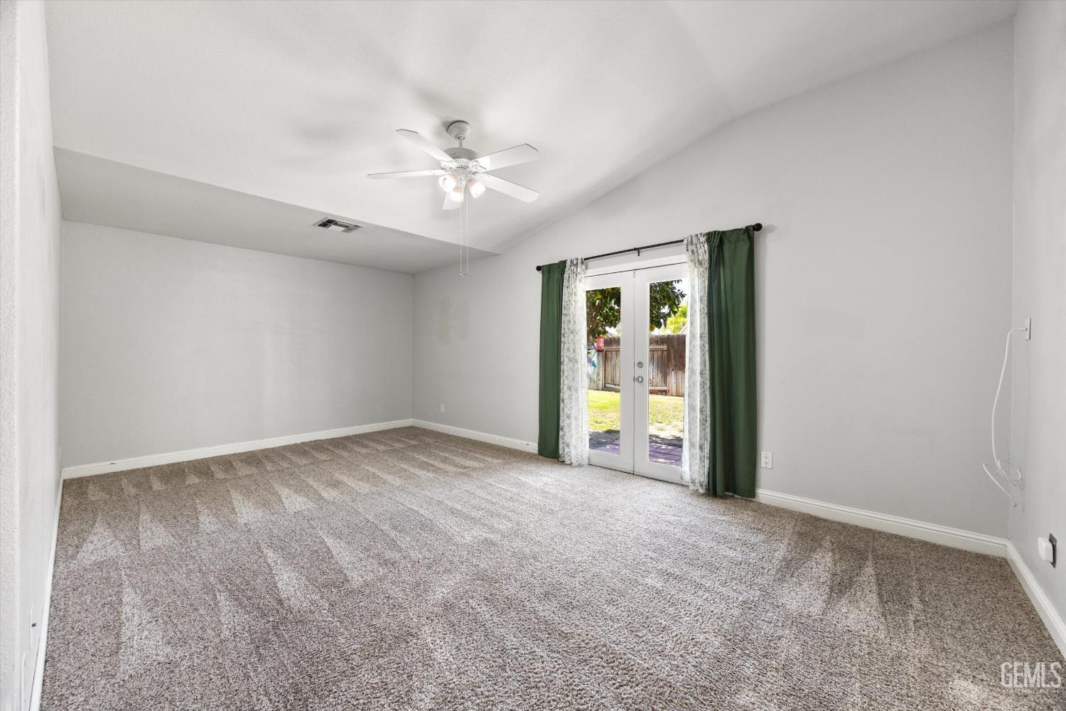 Empty carpeted room with a ceiling fan and glass doors opening to a fenced backyard.