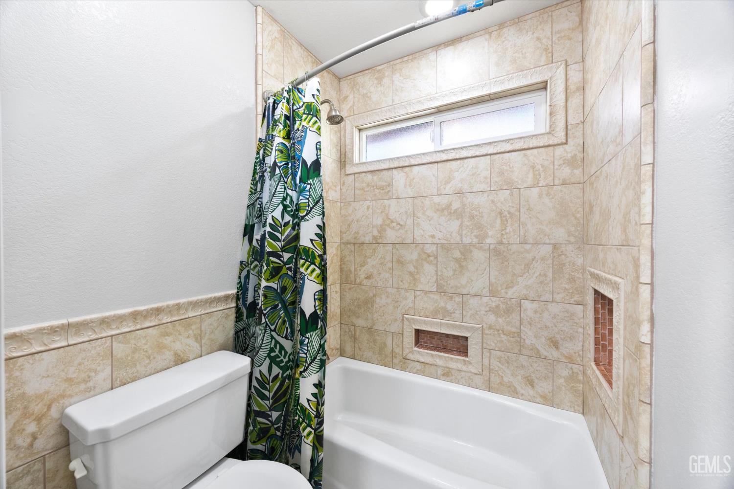 Bathroom with a beige tiled tub surround, built-in niches, and a green leaf-patterned shower curtain.