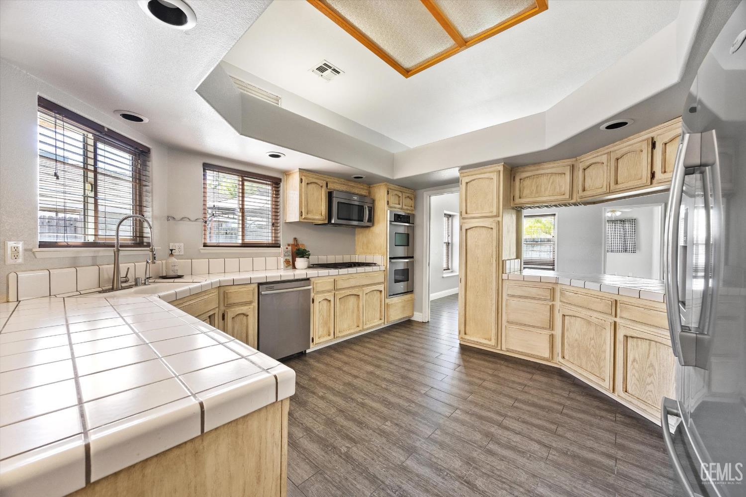 Kitchen with light wood cabinets, tiled counters, stainless appliances, and a wood-trimmed skylight.