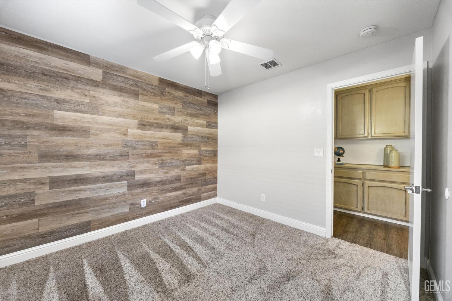 Empty bedroom with a wood-look accent wall, gray carpet, and ceiling fan; built-in cabinetry is visible through the doorway.