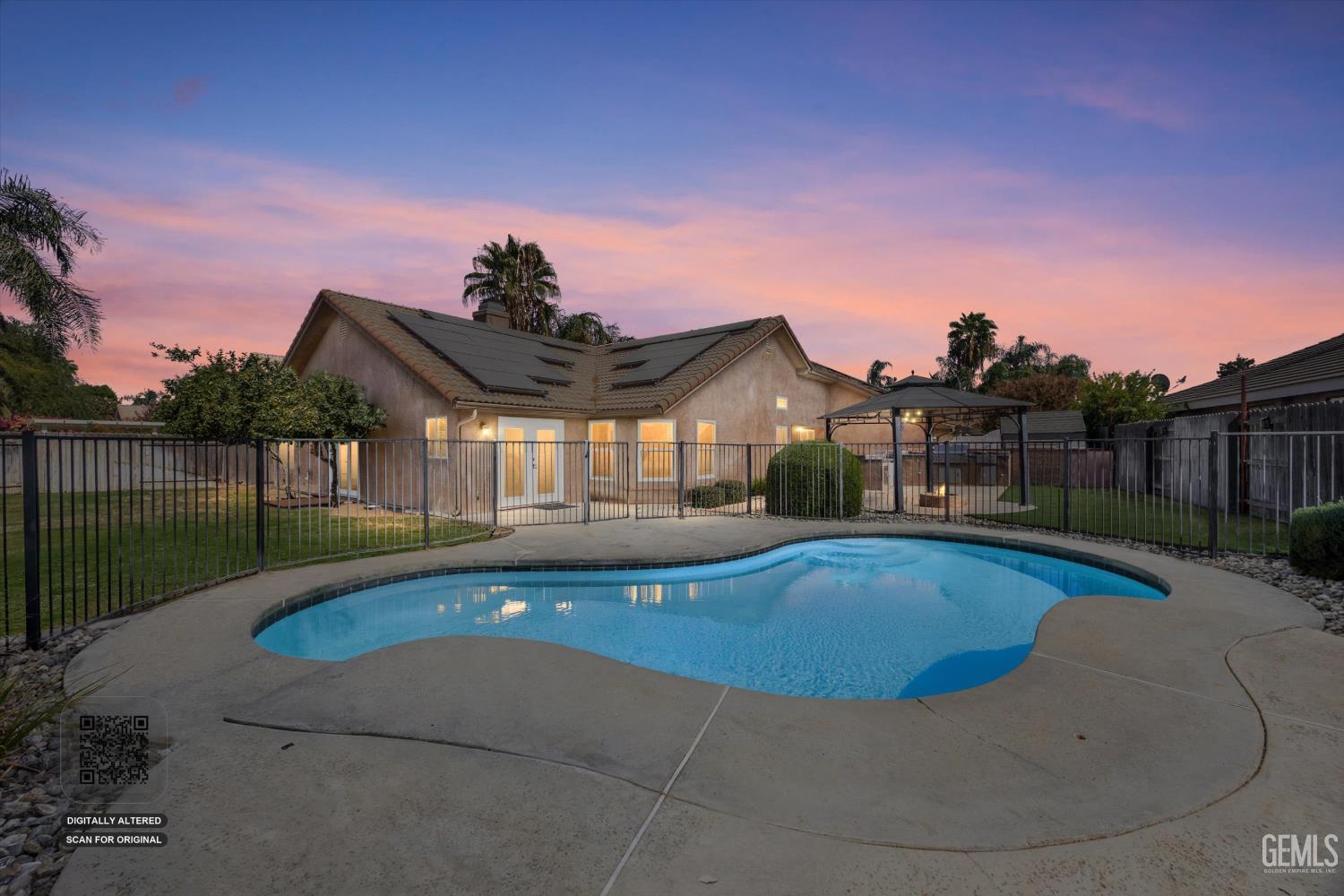 Rear of a single-story home with rooftop solar panels and a freeform pool in a fenced backyard at dusk.