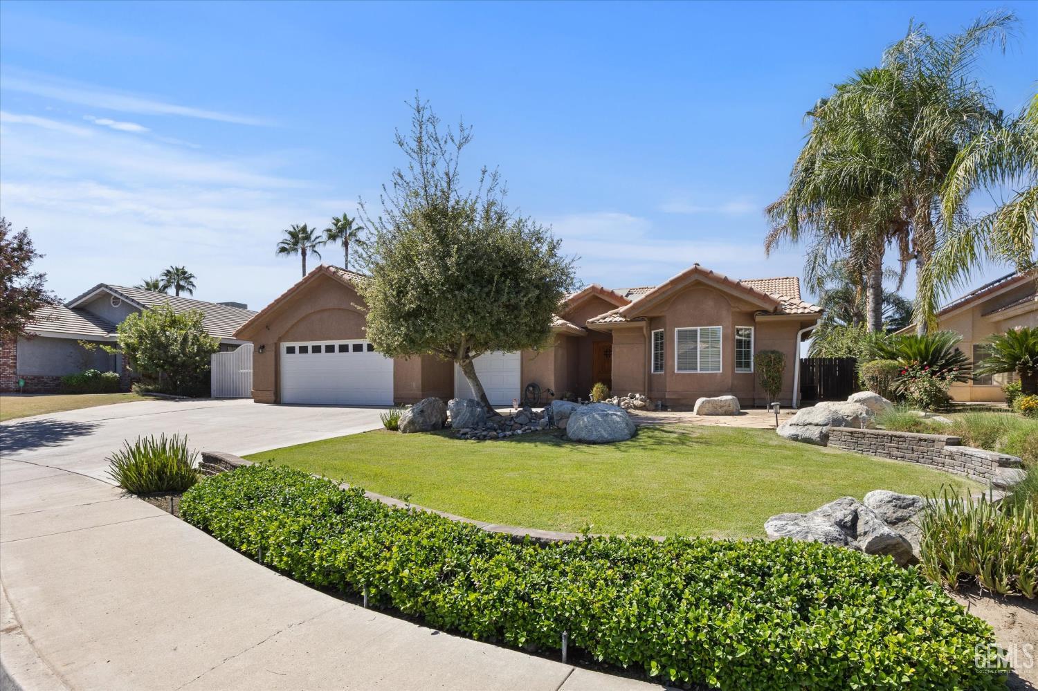 Tan stucco home with tile roof, two-car garage, landscaped lawn, decorative rocks, and palm trees.