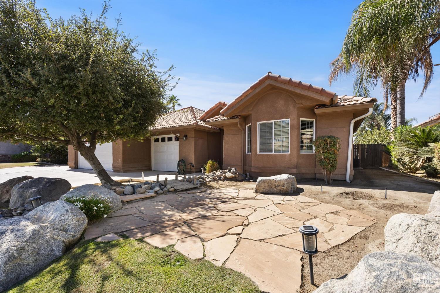 Single-story stucco home with white garage doors and a flagstone path bordered by rocks and trees.