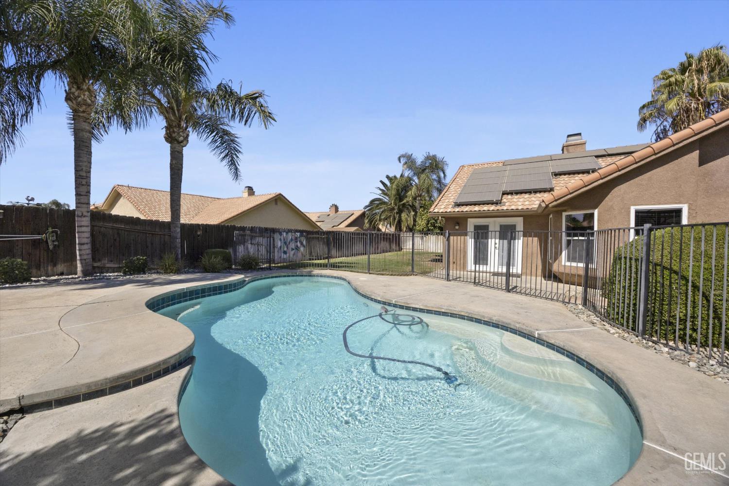 Backyard swimming pool beside a fenced patio and home with rooftop solar panels.