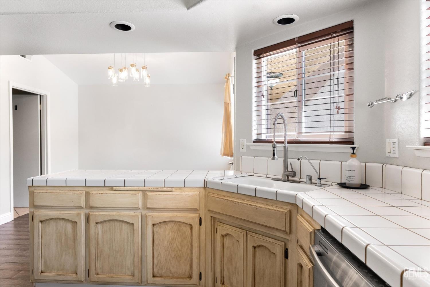 Kitchen with white tile counters, light wood cabinets, and a large window with wooden blinds.