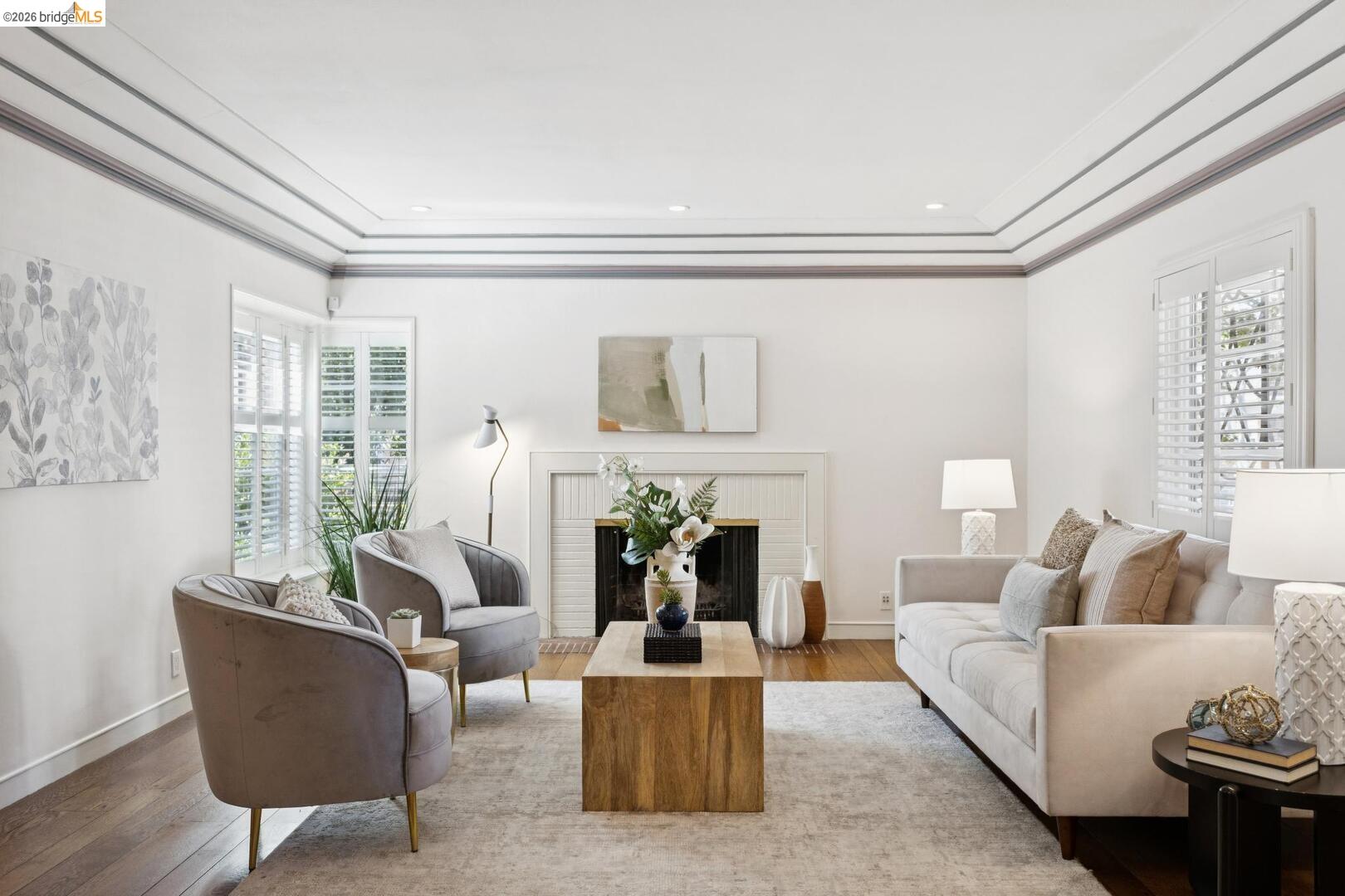 Staged living room with a white fireplace, coffered ceiling, and plantation shutters.