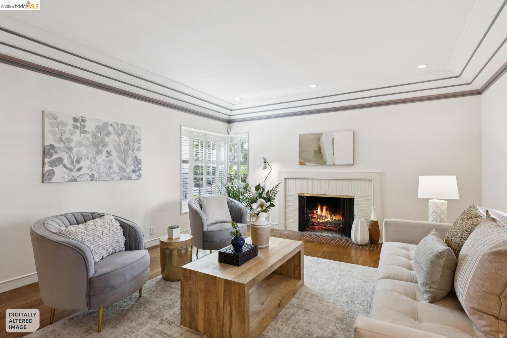 Staged living room with a lit fireplace, shuttered windows, gray armchairs, and a cream sofa.
