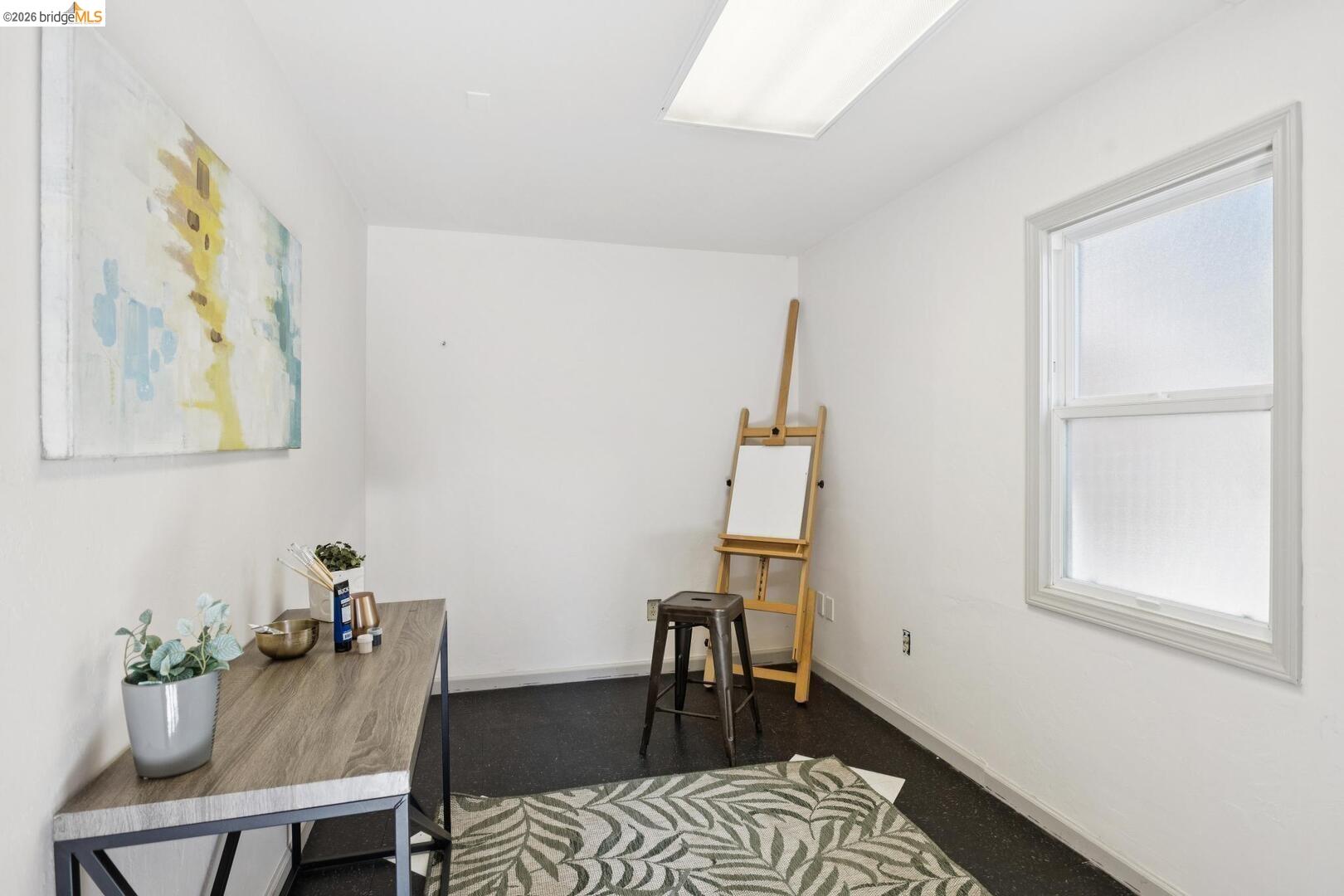 Home office with an easel, desk, large window, and patterned rug.