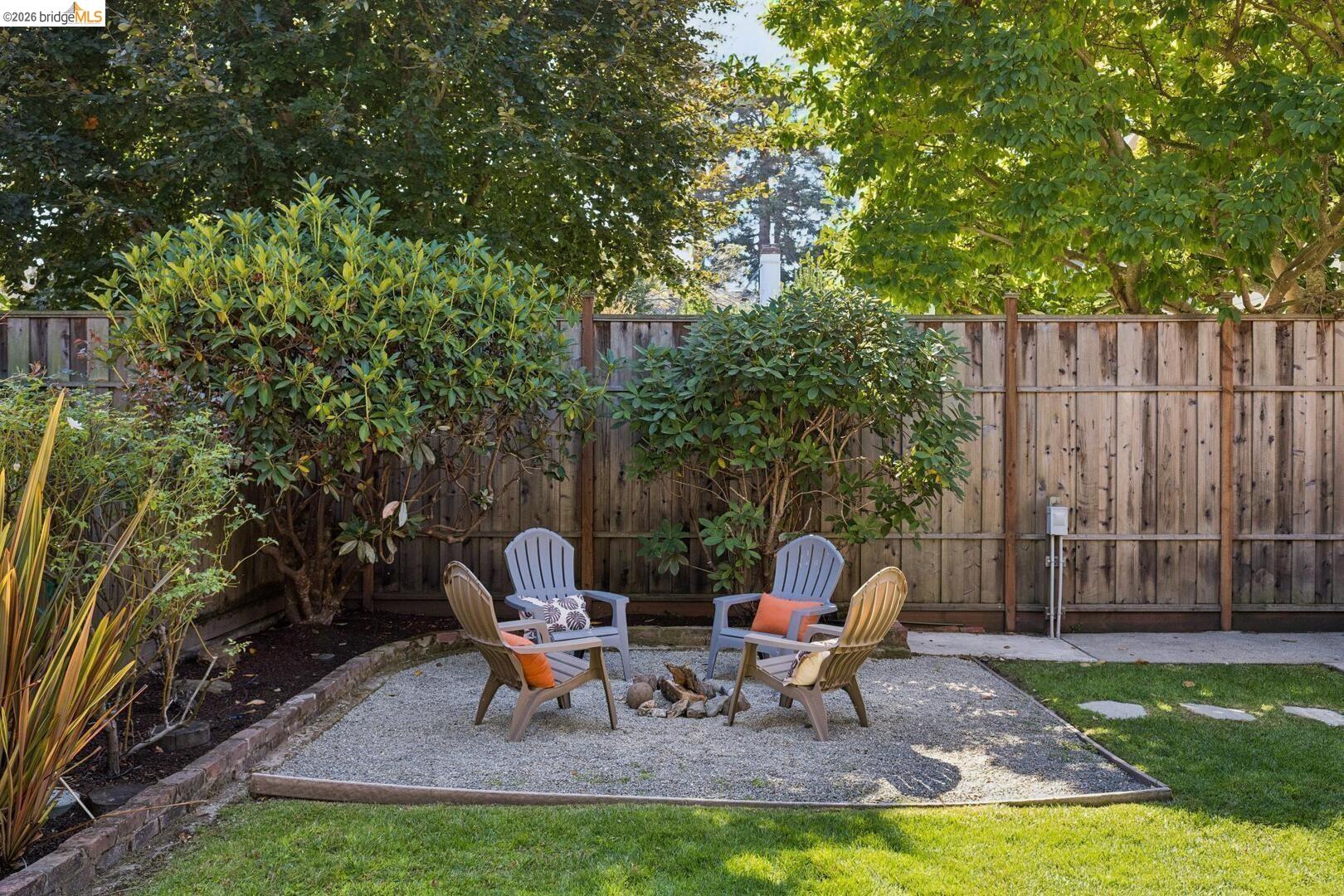 Backyard gravel patio with chairs around a fire pit, bordered by a wooden fence and mature trees.