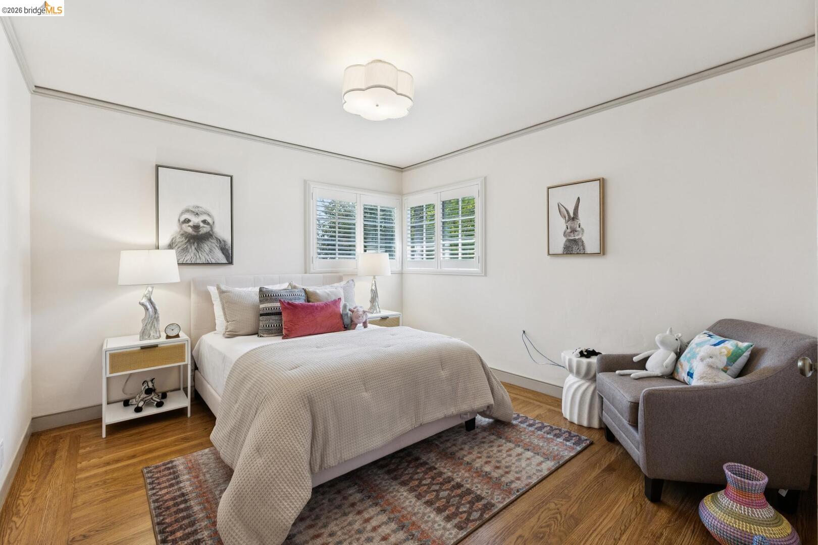 Staged bedroom with a white bed, shuttered corner windows, hardwood floors, and a gray armchair.