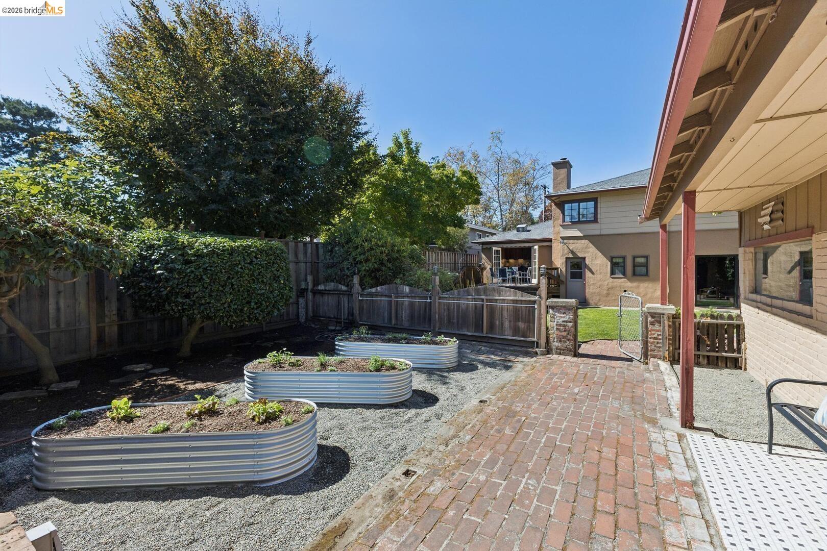 Backyard courtyard with raised garden beds, brick walkway, fenced lawn, and covered patio.