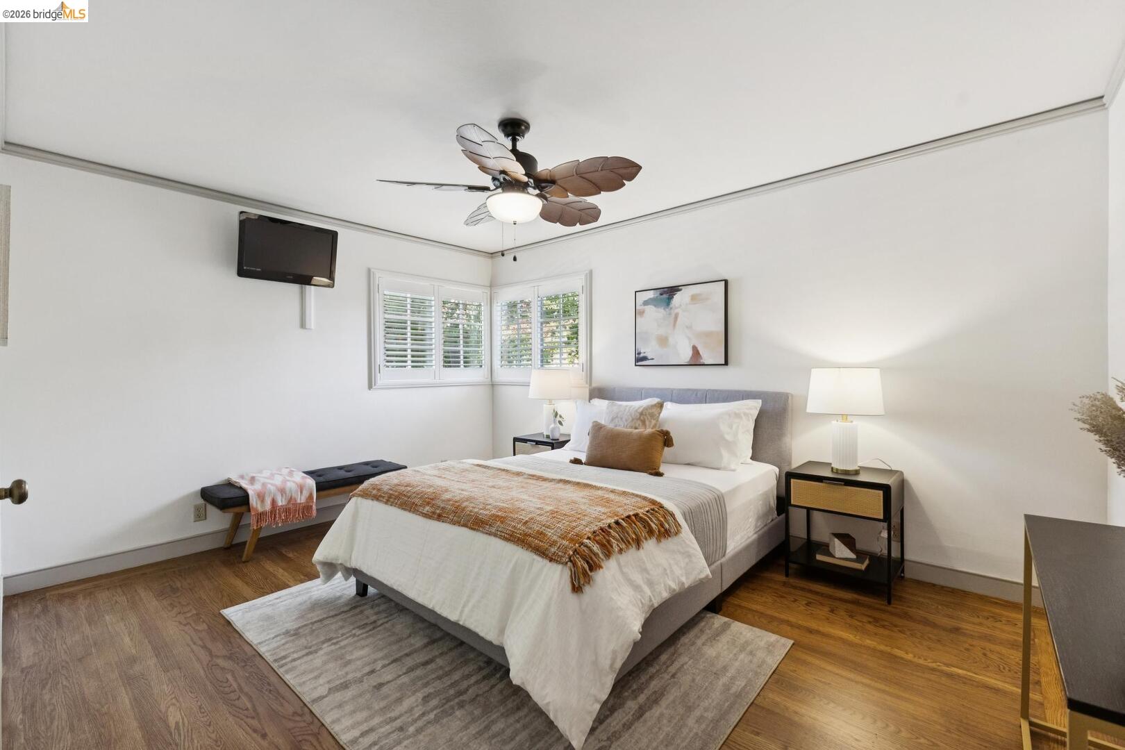 Staged bedroom with a gray upholstered bed, hardwood floors, and plantation shutters on corner windows.