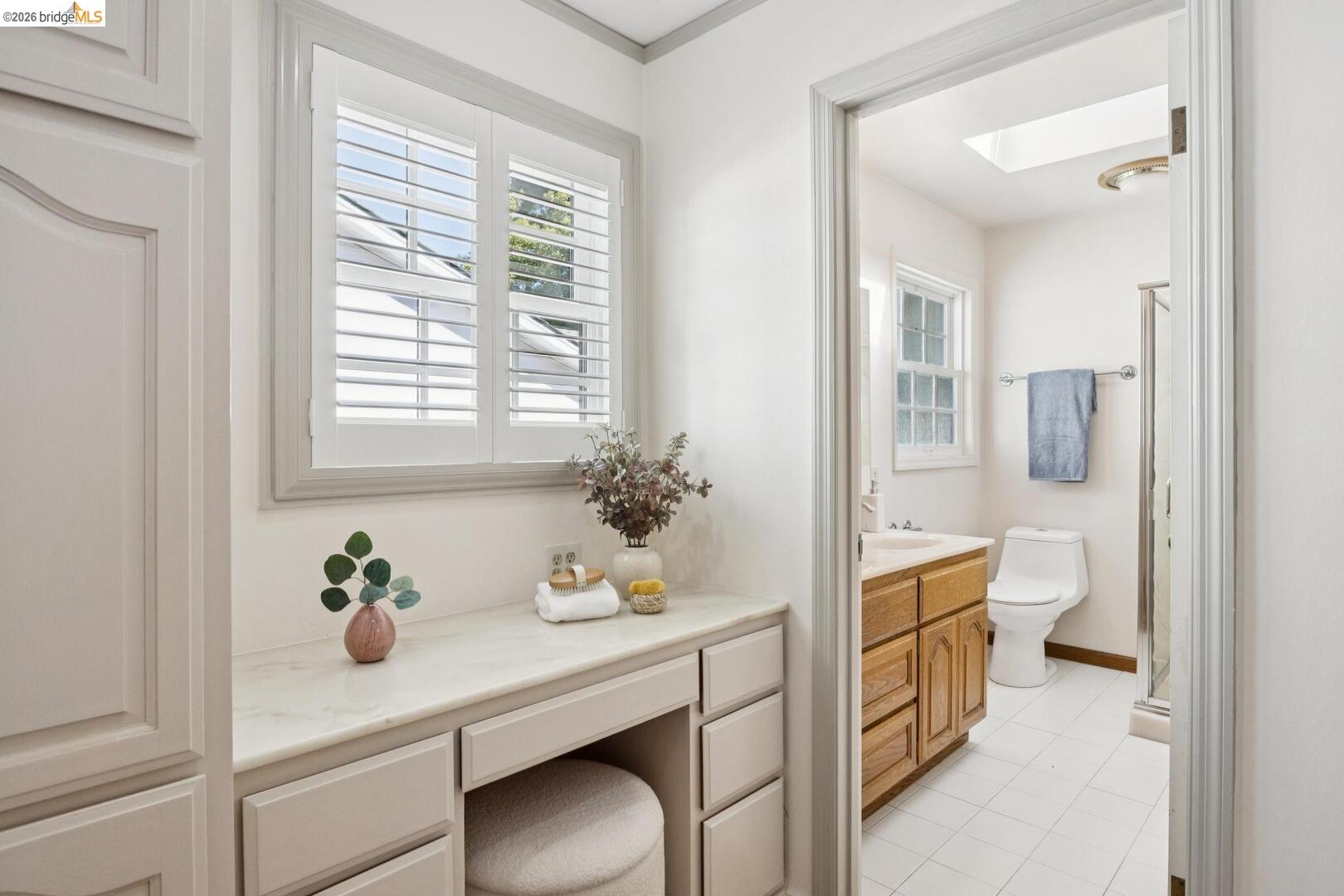 Bathroom vanity with shuttered window, opening to a second vanity, toilet, and glass shower.
