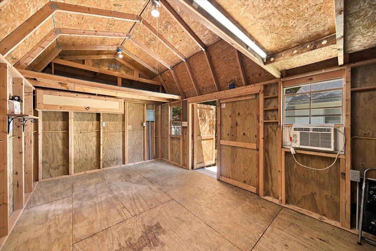 Unfinished garage with exposed wood framing, vaulted ceiling, plywood floor, and a window air conditioner.