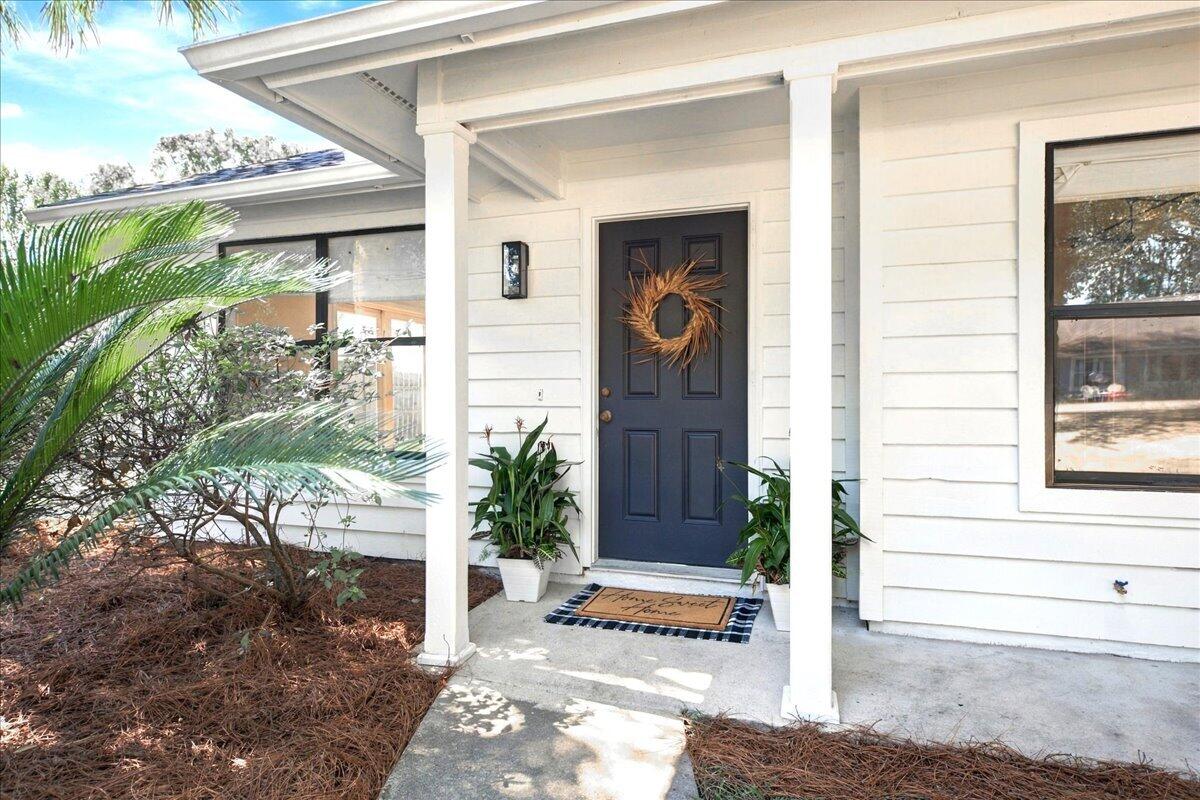 White house entrance with a covered porch, dark blue front door, wreath, and potted plants.
