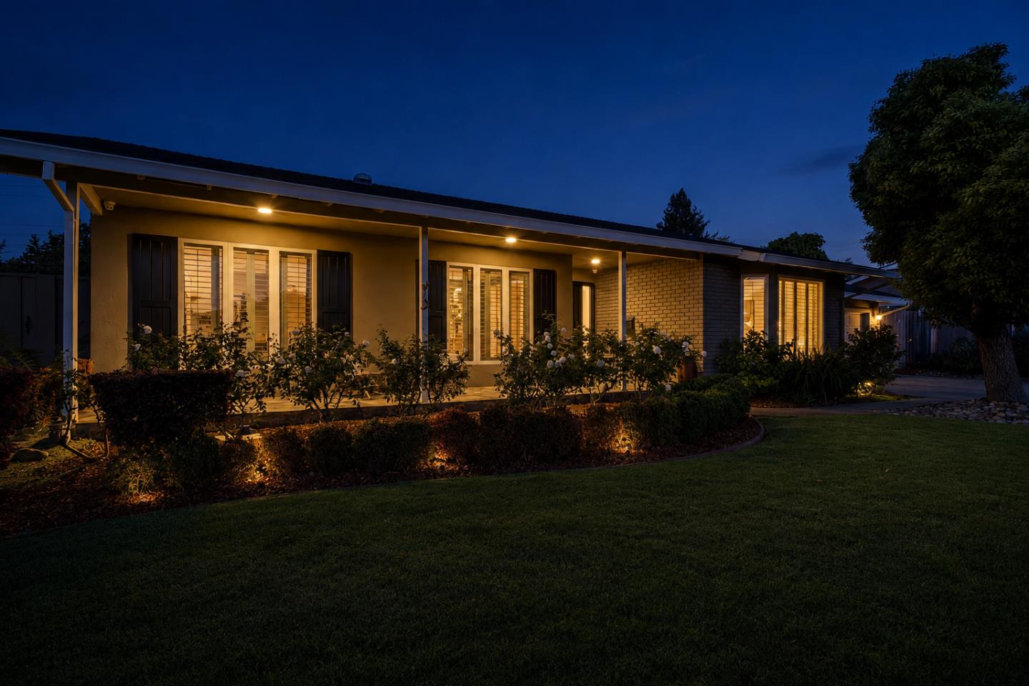 Single-story home at dusk with a covered porch, glowing windows, landscaped flower beds, and front lawn.