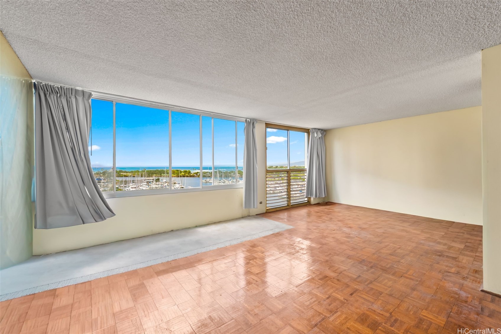 Vacant living room with parquet floors and broad windows overlooking a marina and ocean.