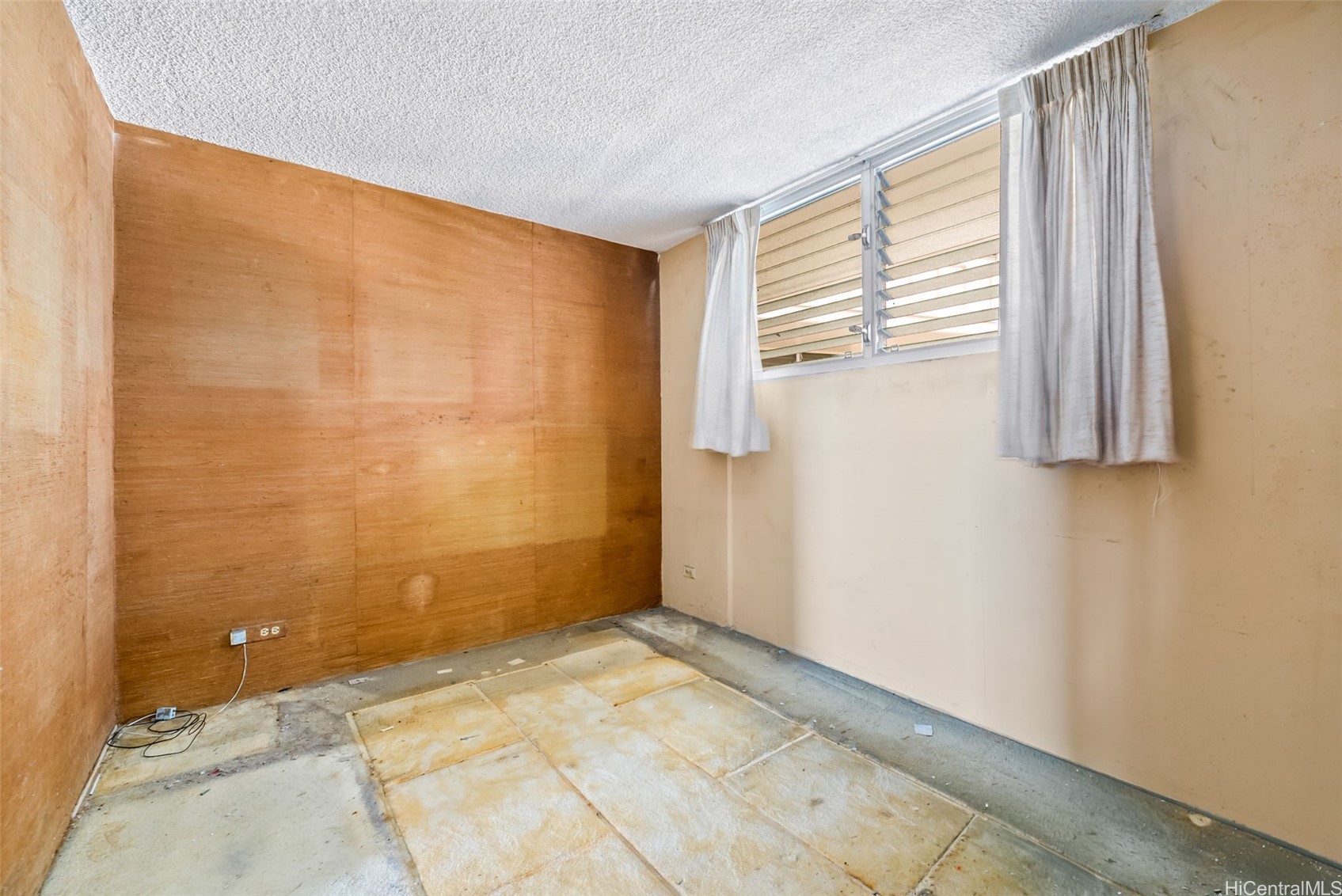 Empty room with wood-paneled walls, a curtained louvered window, and an unfinished floor.