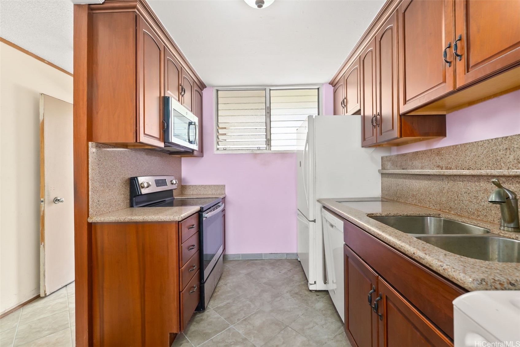 Galley kitchen with reddish wood cabinets, speckled counters, white appliances, and a louvered window.