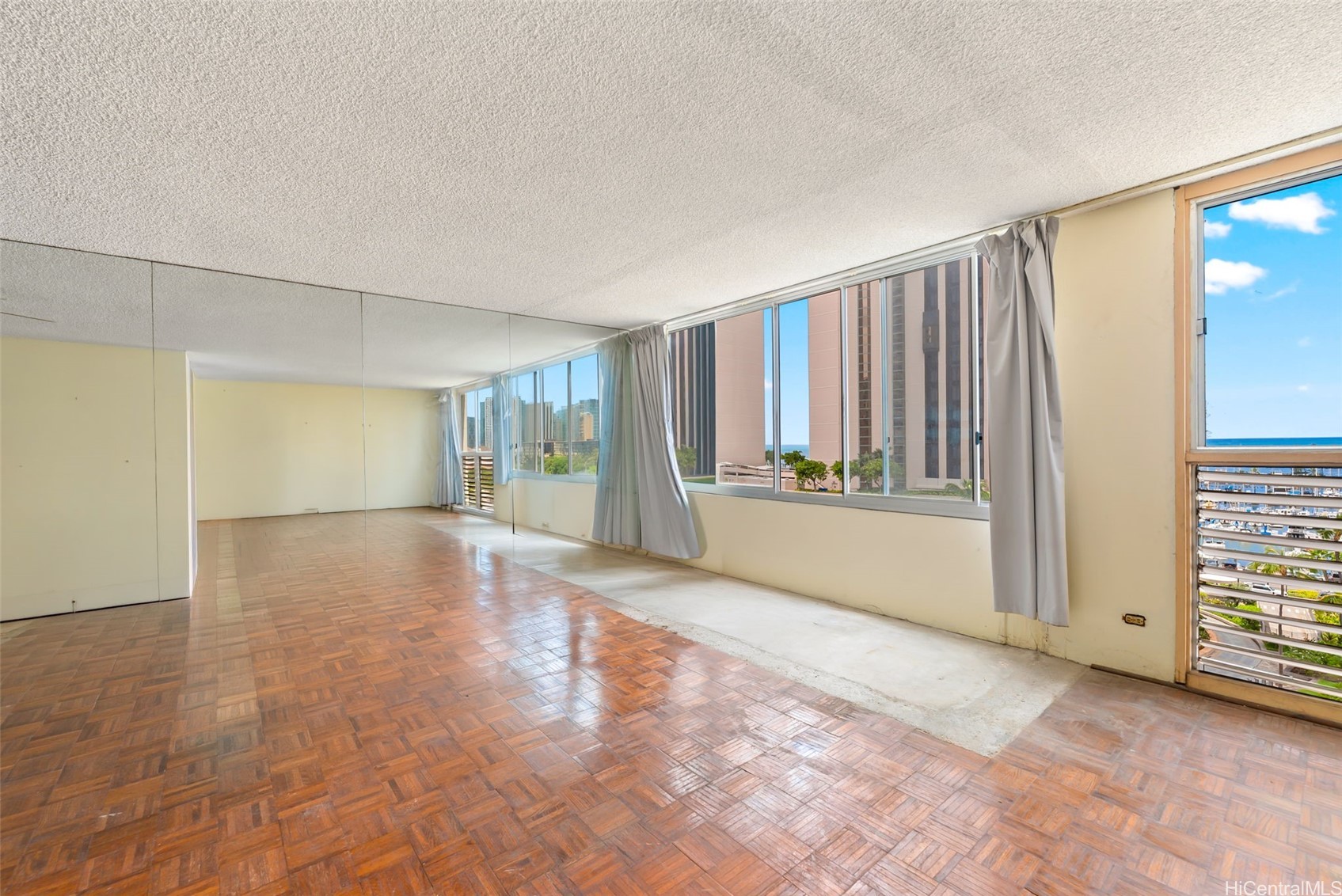 Empty living room with mirrored walls, parquet floors, and windows overlooking high-rises and the ocean.