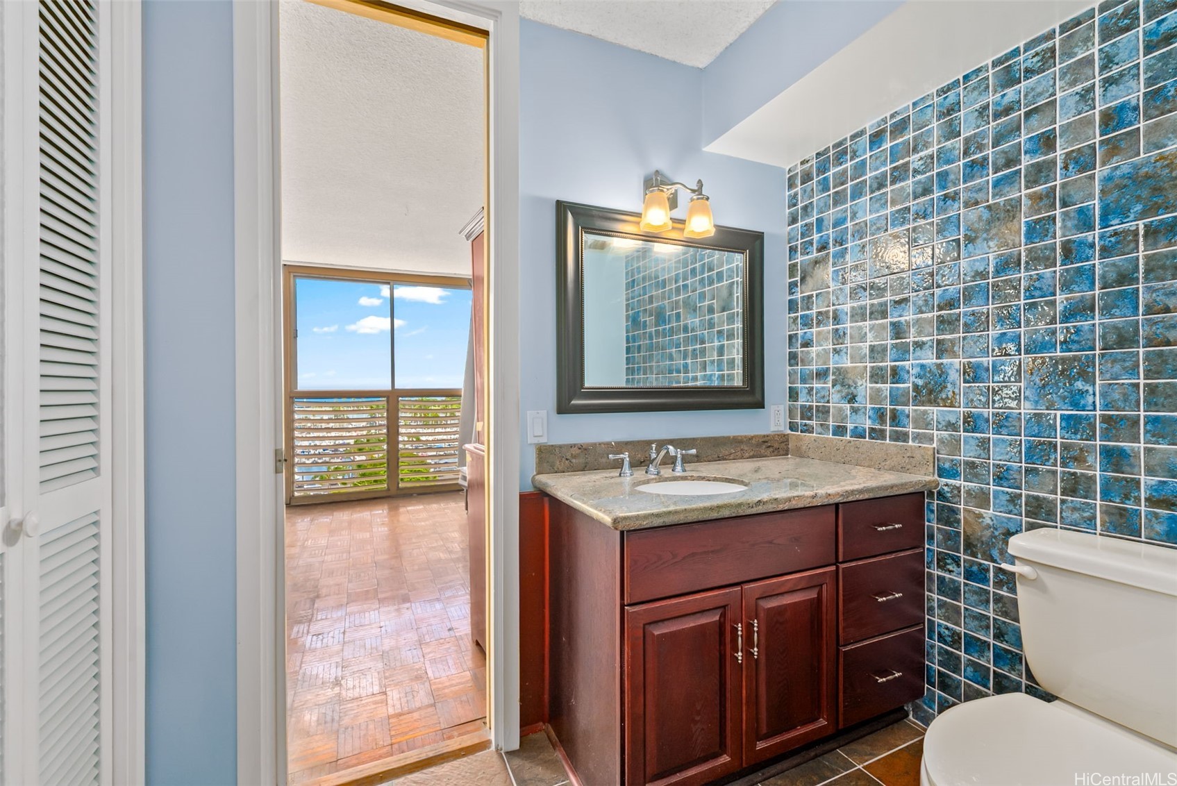 Bathroom with blue mosaic tile, dark wood vanity, and doorway to a bright room with a balcony.