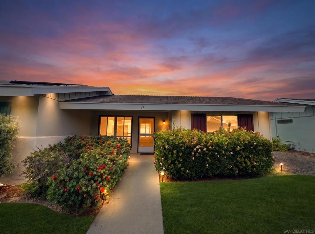 Single-story home with a lit entry and flowering shrubs beneath a colorful dusk sky.