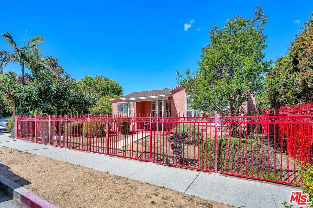 Single-story home behind an ornate red metal fence, with mature trees and a front walkway.