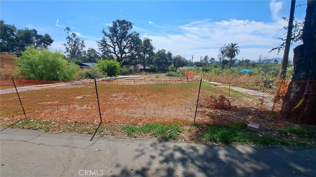 Vacant fenced lot with dry ground and mature trees, viewed from the sidewalk.