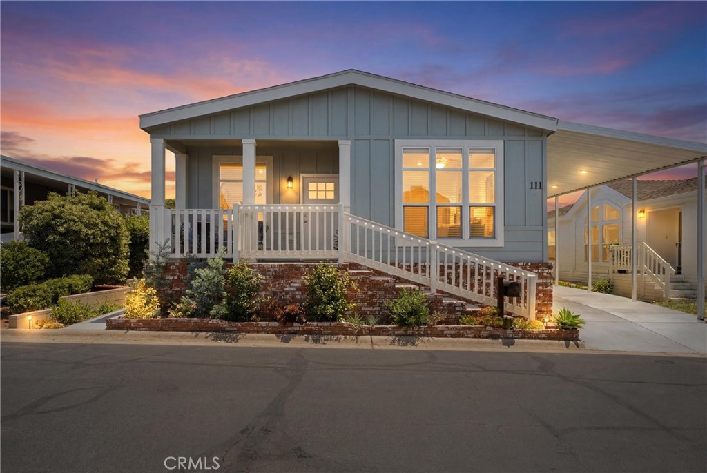 Gray home with a covered porch and landscaped flower beds at dusk.
