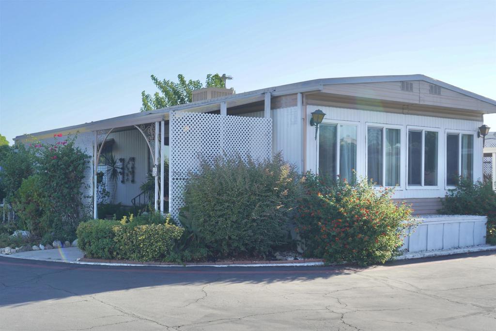 Single-story manufactured home with a covered entrance, white lattice panels, and flowering shrubs.