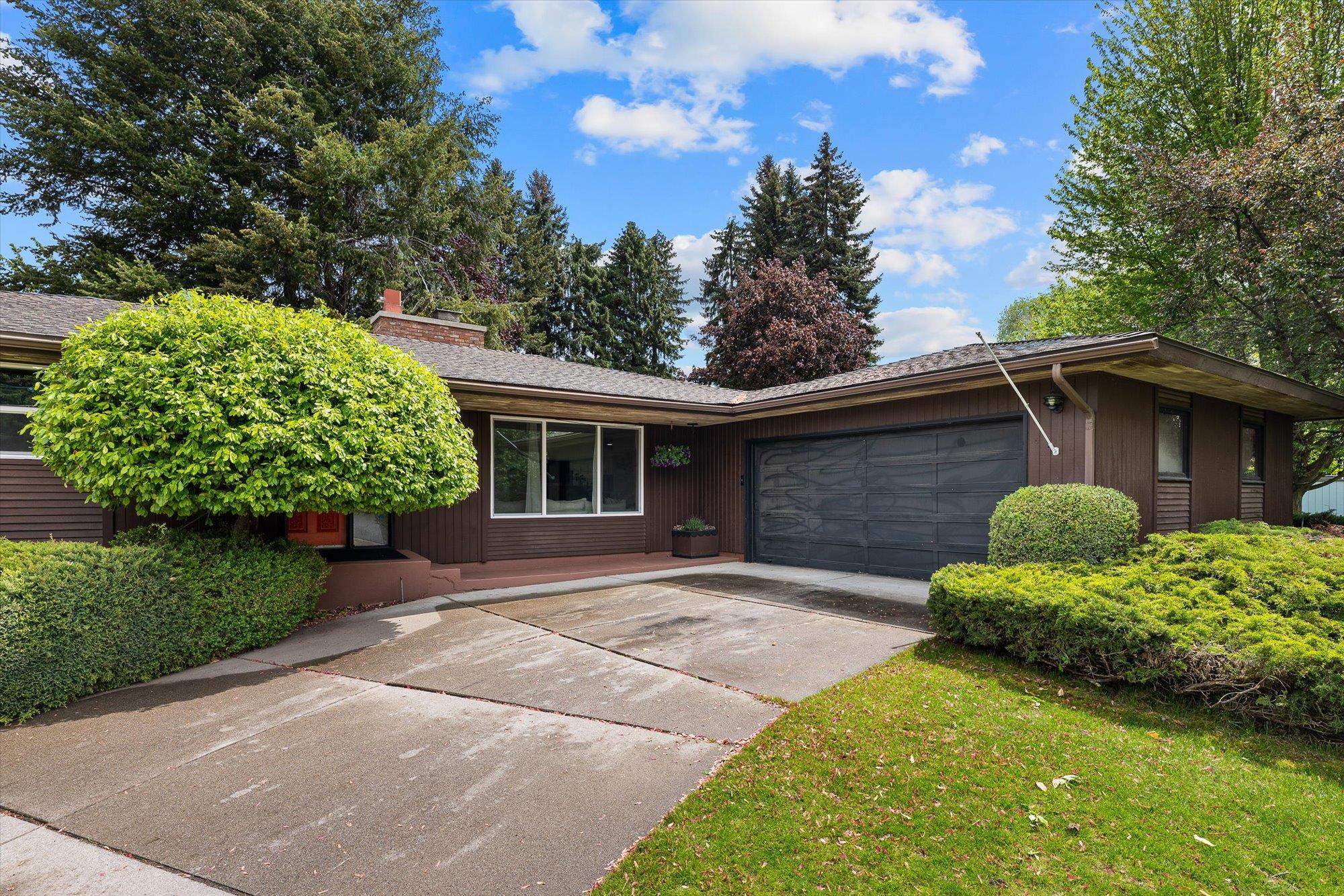 Single-story brown ranch house with an attached garage, wide driveway, and mature landscaping.