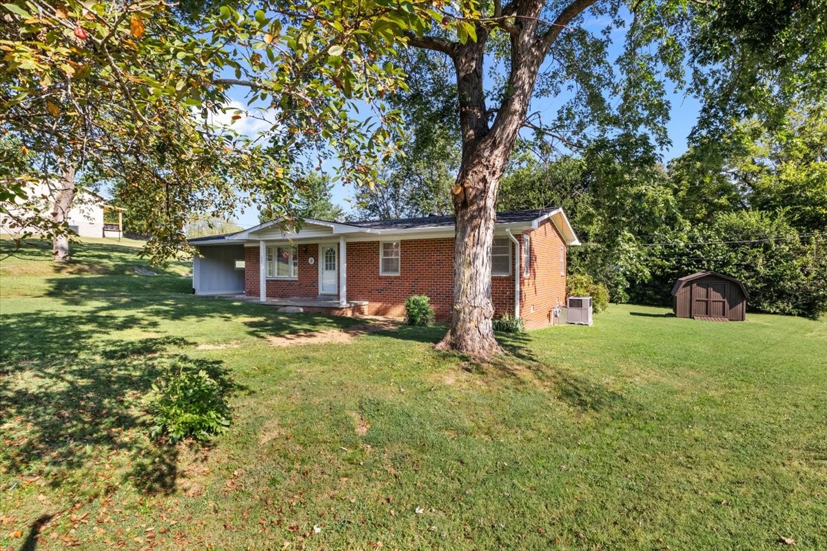 Single-story brick home with a carport, mature trees, and a small shed across the lawn.