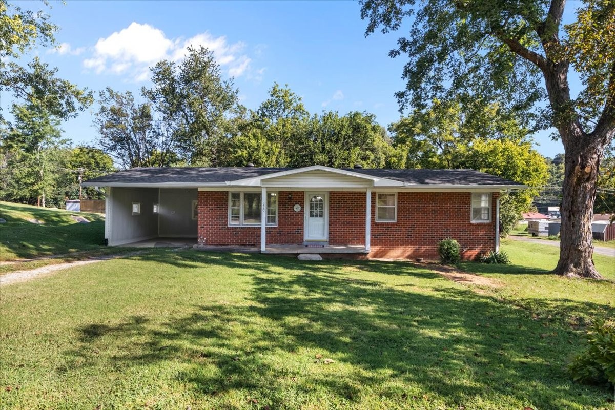 Red-brick ranch house with a covered porch and attached carport beneath mature trees.