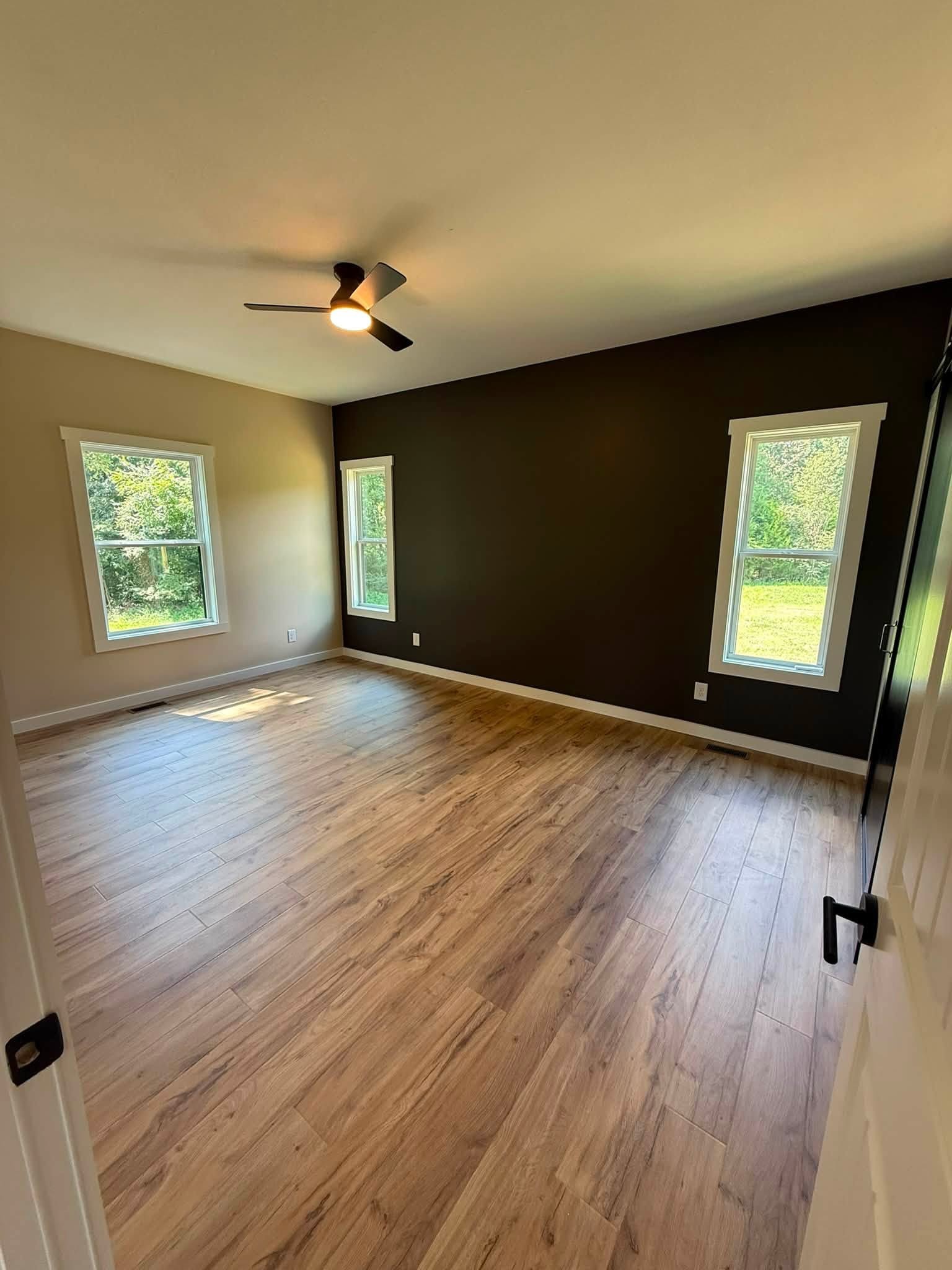 Empty bedroom with wood-look flooring, a dark accent wall, three windows, and a ceiling fan.