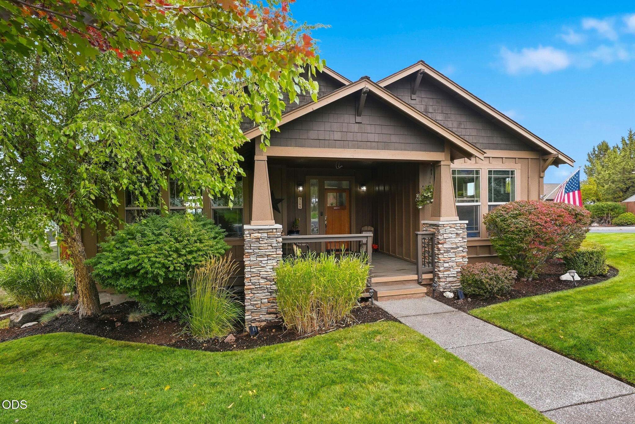 Brown shingle-sided home with a covered porch, stone columns, and landscaped front lawn.