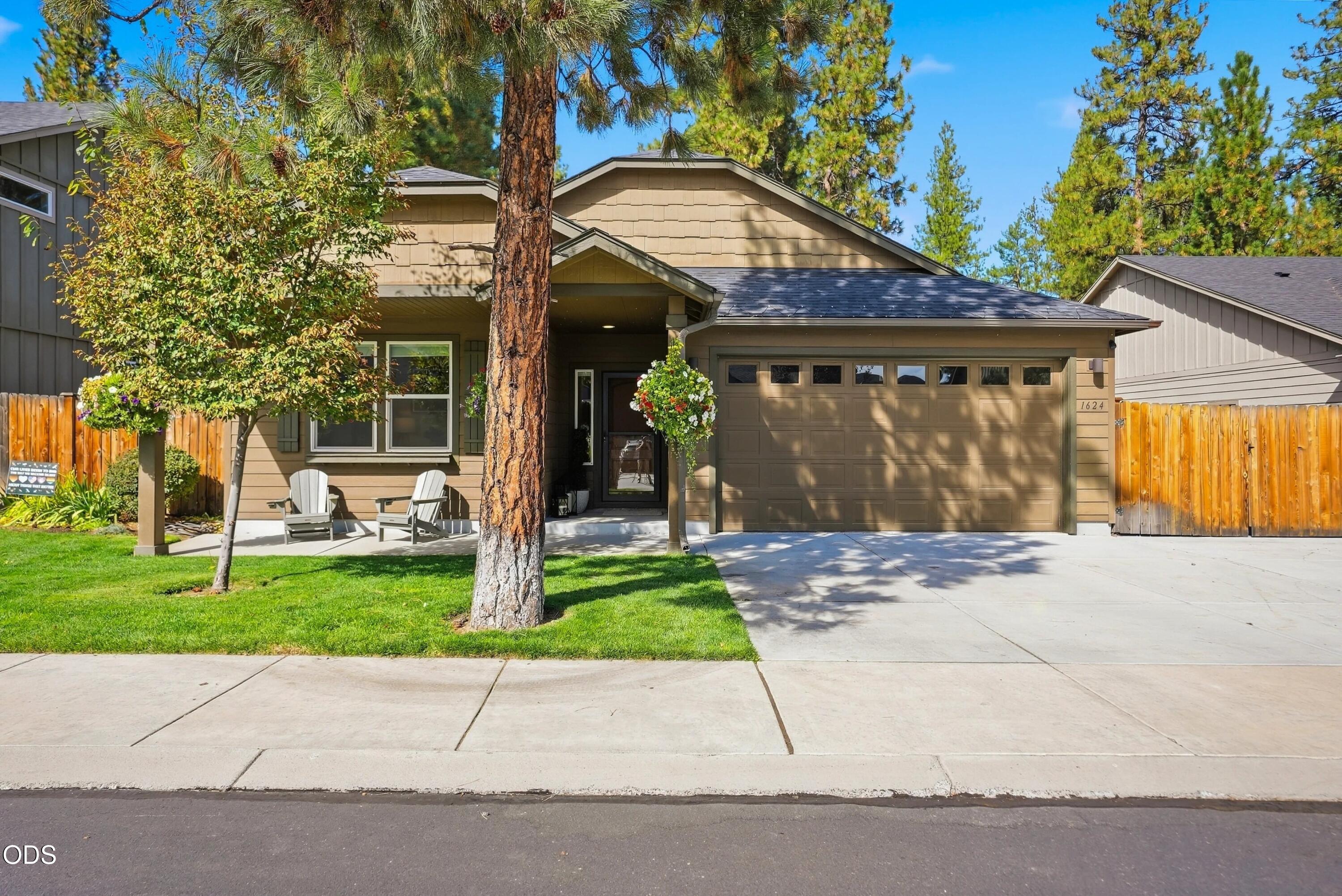 Tan single-story home with an attached garage, shaded front porch, green lawn, and mature trees.