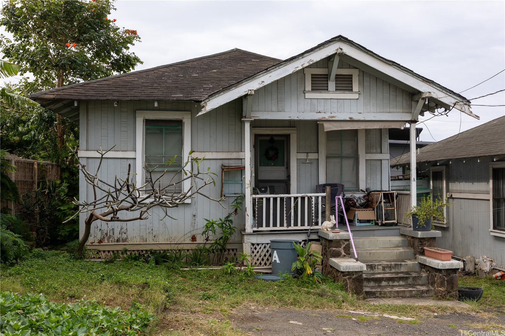 Weathered gray wooden house with a front porch, concrete steps, and overgrown yard.