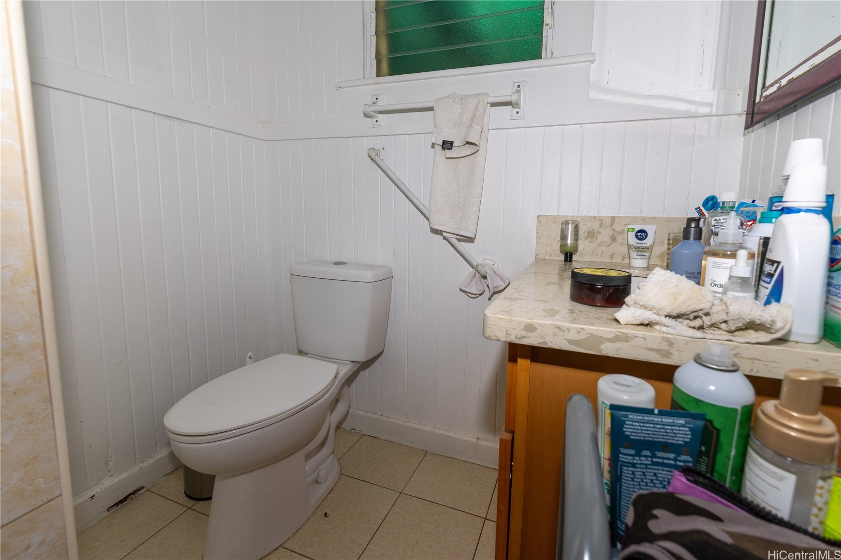 Bathroom with a white toilet, tiled floor, and marble-look vanity crowded with toiletries.