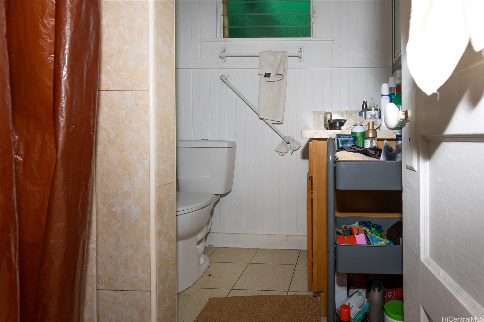 Compact bathroom with a toilet, beige-tiled shower, brown curtain, and crowded storage shelves.