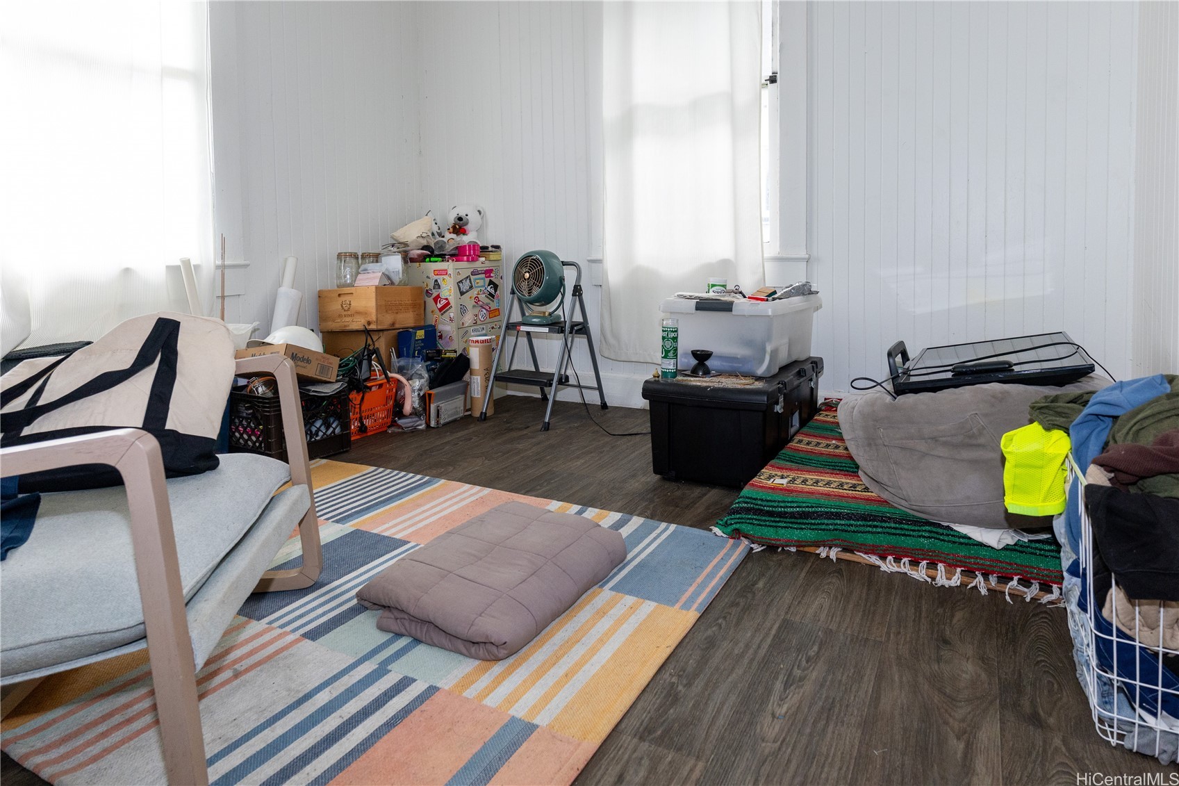 Cluttered room with white paneled walls, tall windows, dark flooring, chairs, rugs, and storage bins.