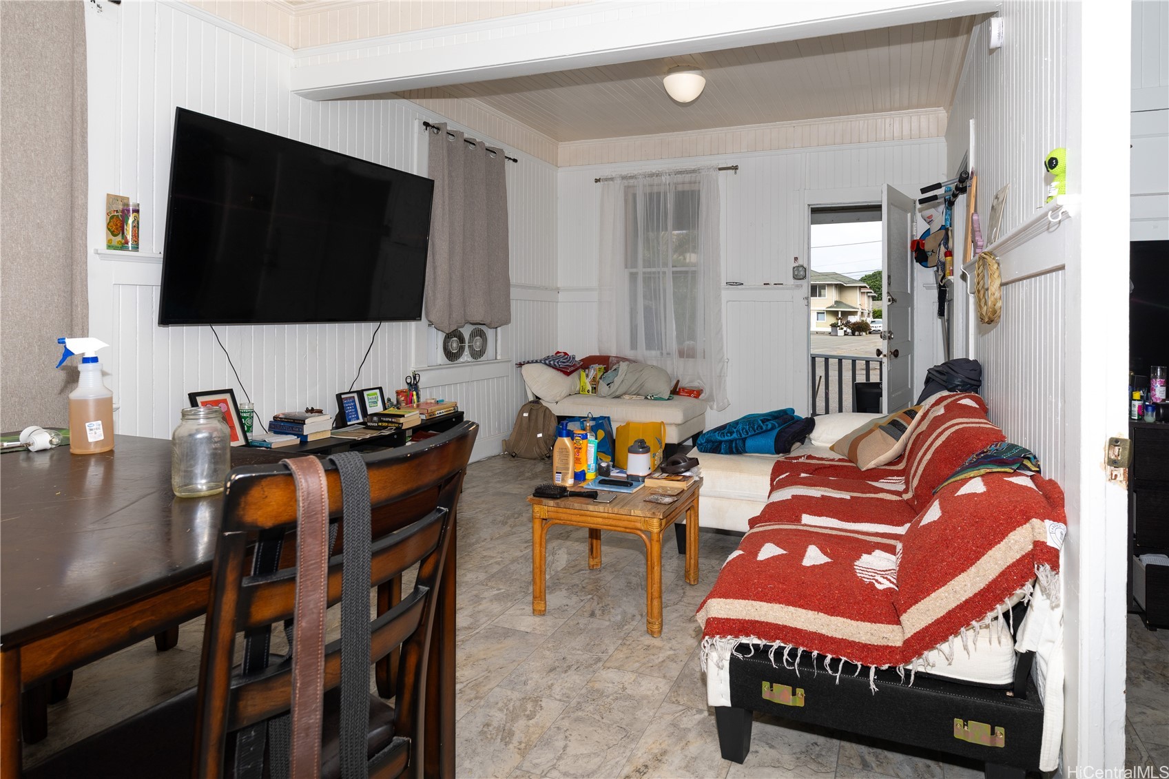Living room with white paneled walls, a large mounted television, sofas, and an open front door.