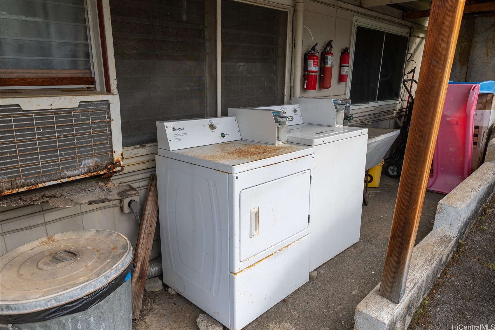 Covered outdoor laundry area with two white appliances and a large metal trash can.