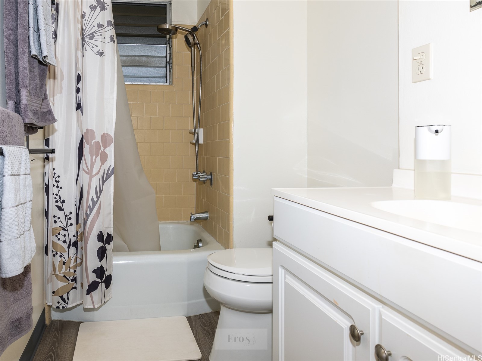 Bathroom with a tiled tub and shower, floral curtain, toilet, and white vanity.