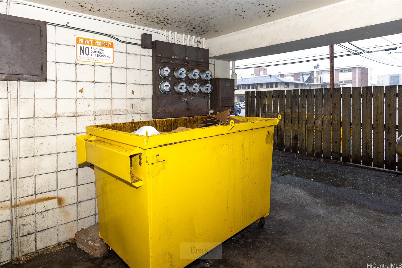 Yellow commercial dumpster in a covered service area beside utility meters and a wooden fence.