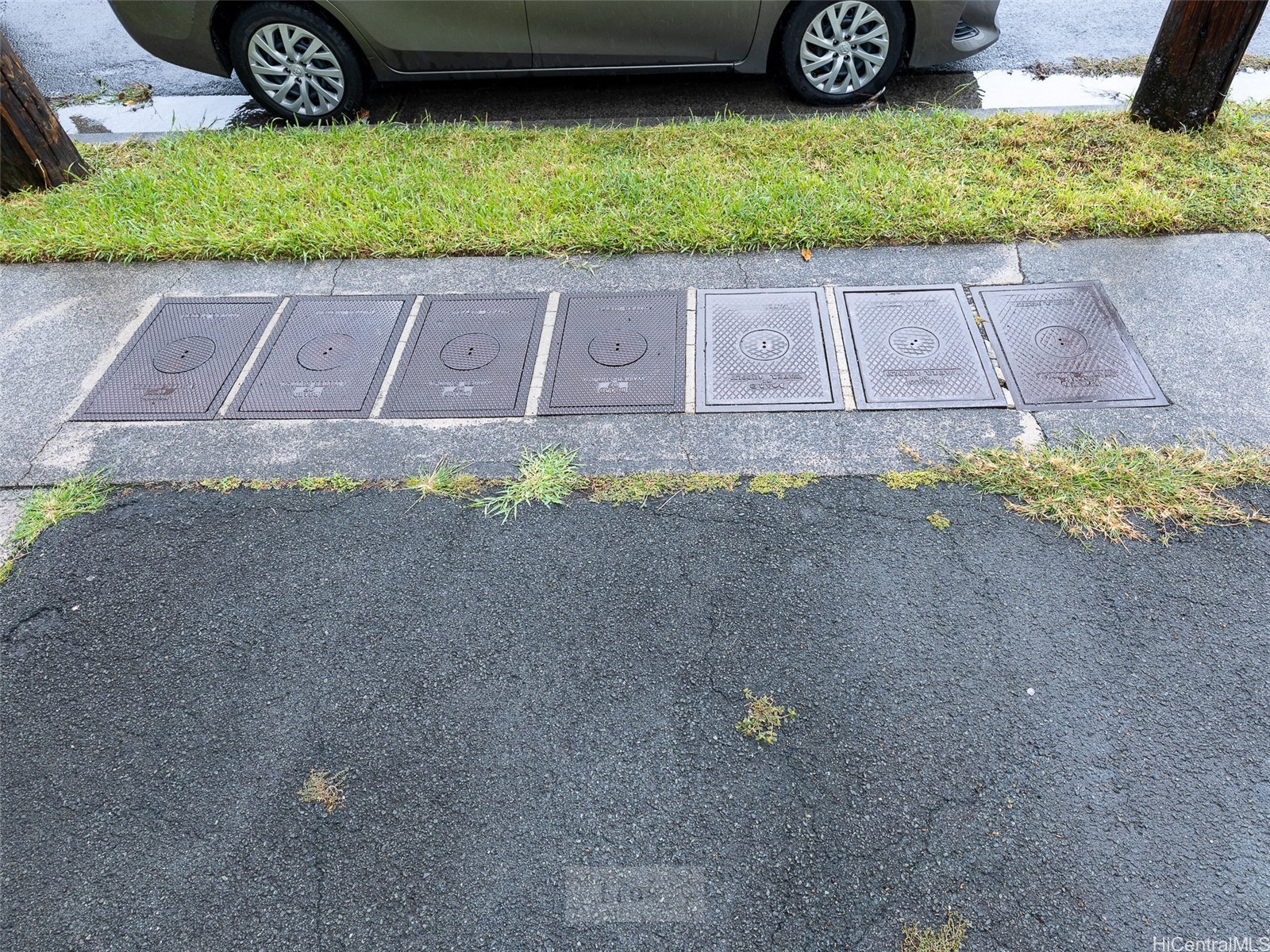 Seven utility access covers set into a sidewalk beside a grassy verge and asphalt road.