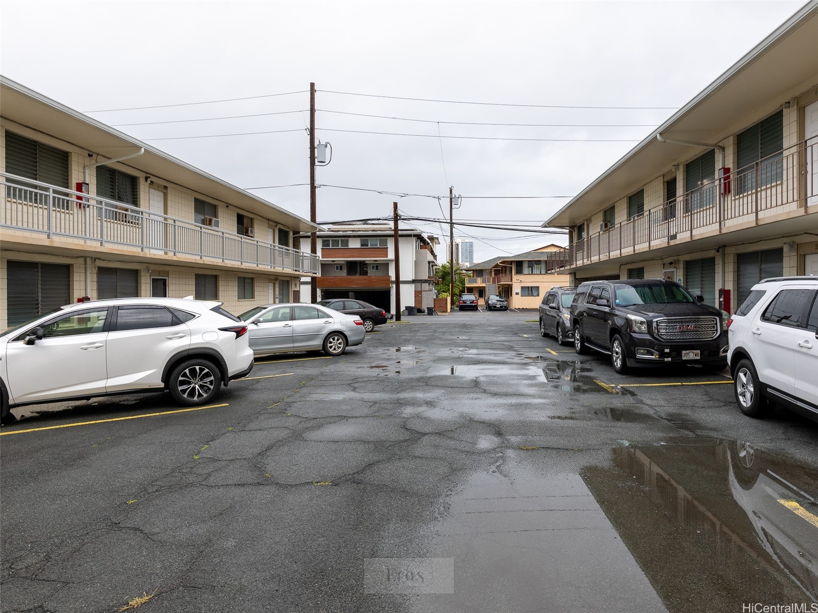 Apartment building parking lot with parked cars and puddles on wet pavement.