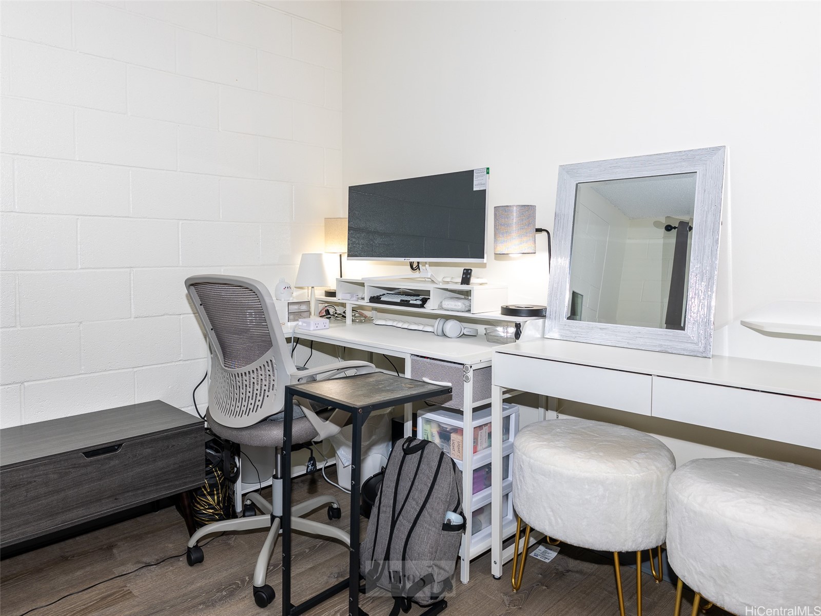 Compact home office with a desk, monitor, ergonomic chair, large mirror, and white upholstered stools.