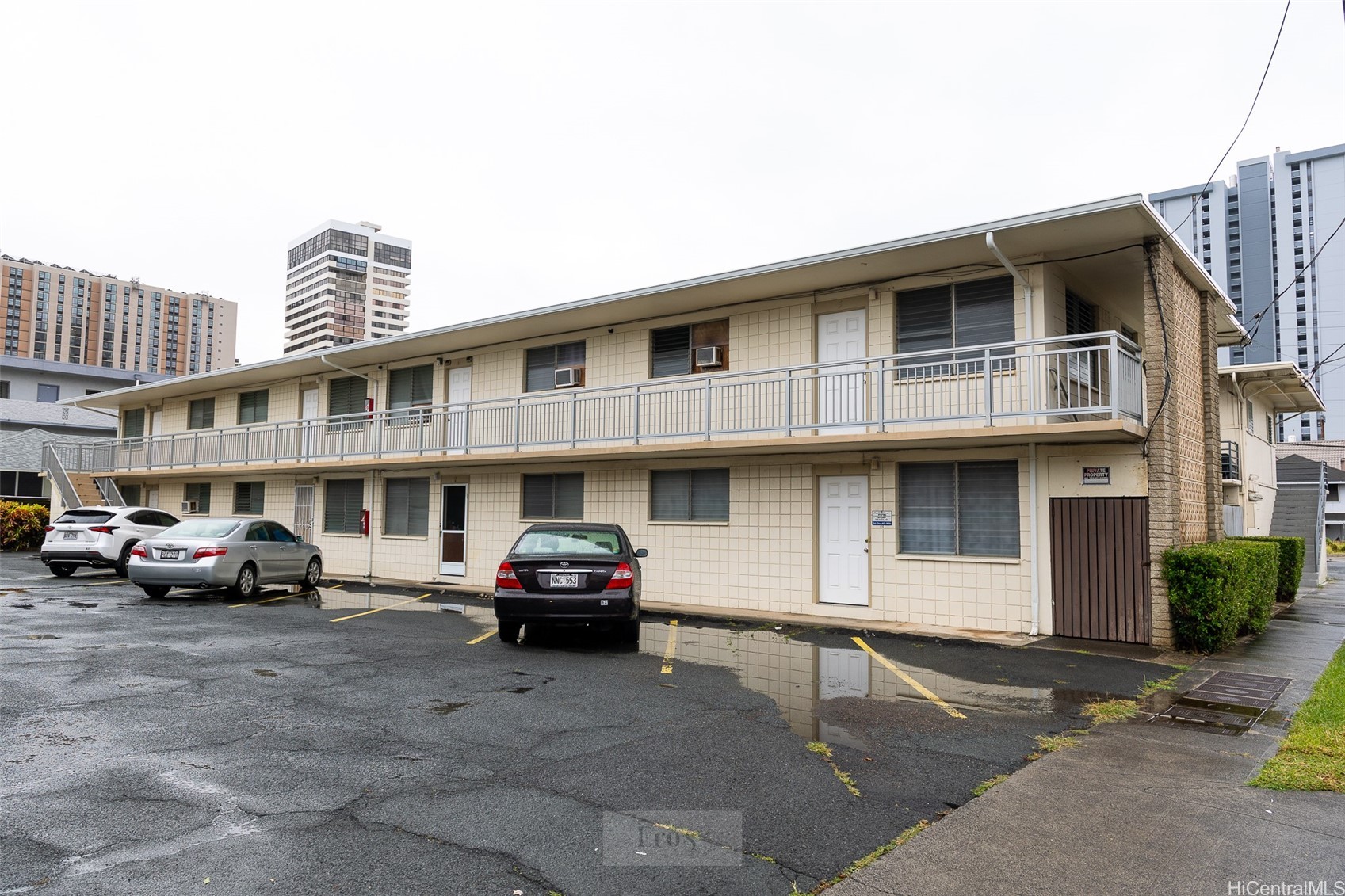 Two-story apartment building with open-air walkways and a wet parking lot in front.